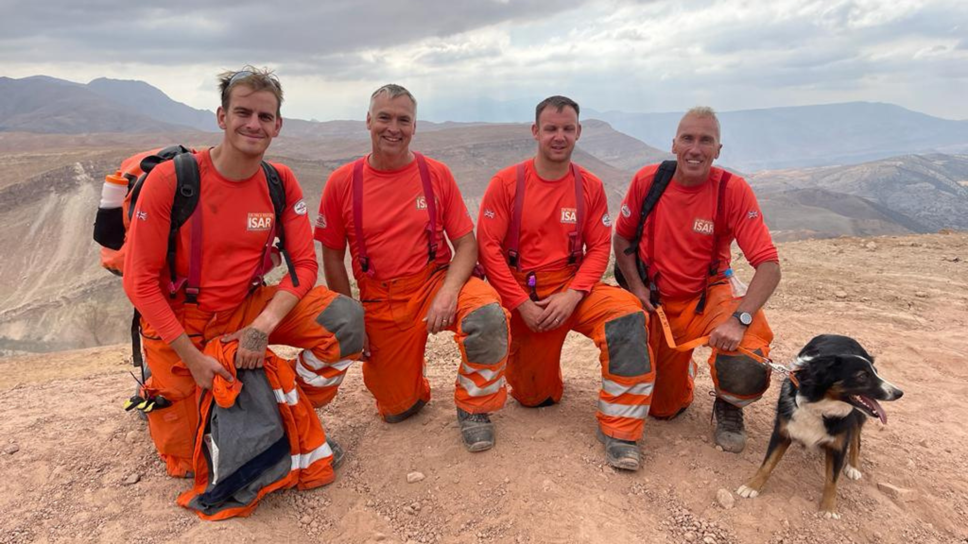 Four firefighters standing in a remote sandy landscape kneeling on one knee with a dog, smiling at the camera
