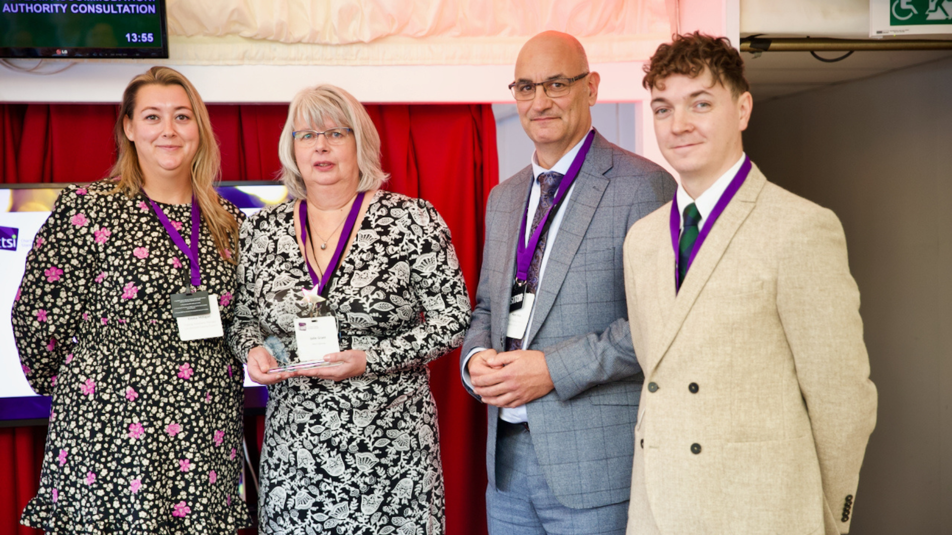 Two women stood sided by side wearing a purple lanyard and two men to the right wearing purple lanyards all wearing formal clothing at an awards ceremony