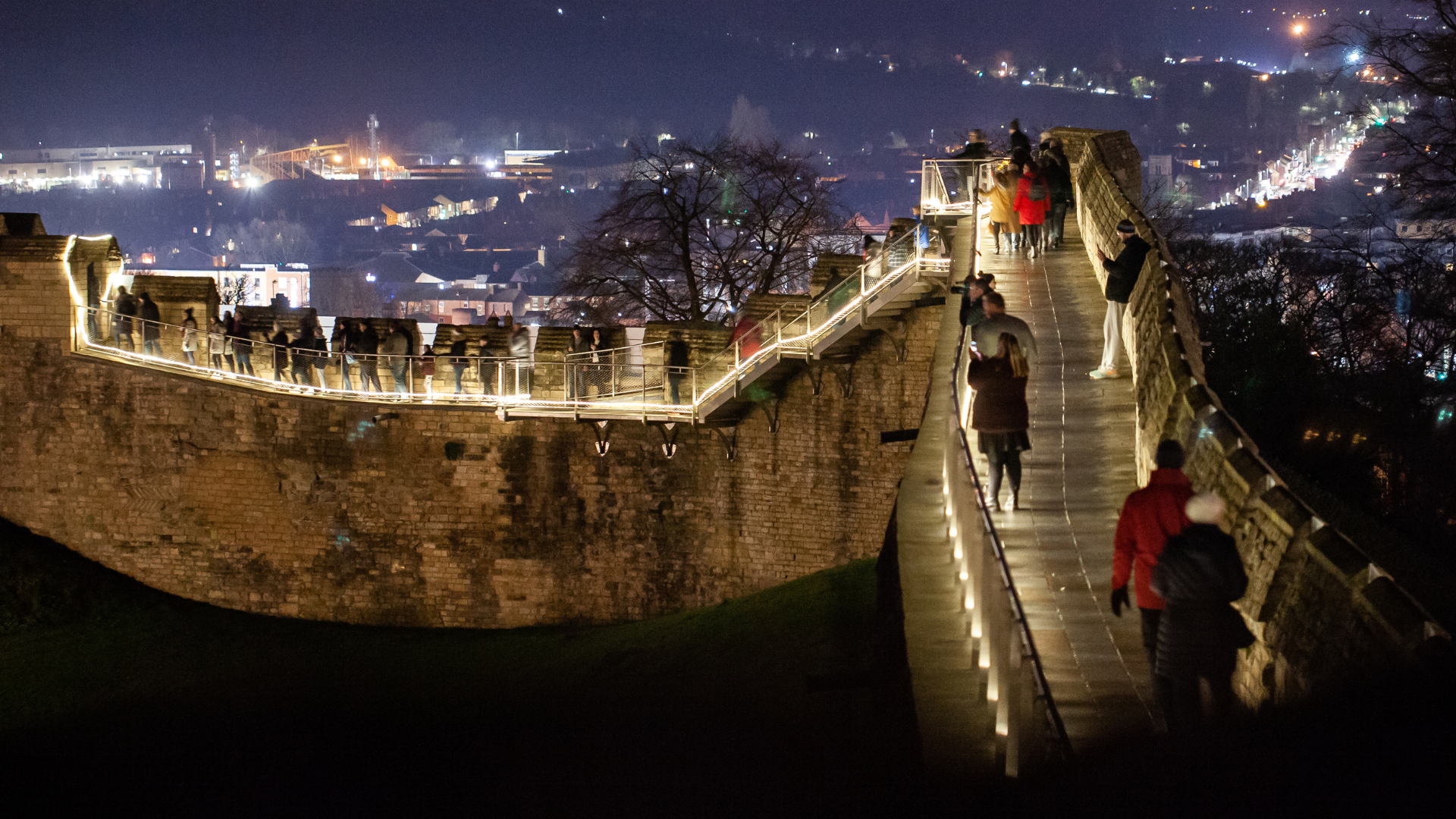 A medieval wall walk illuminated by lights at dark, with people walking along the wall