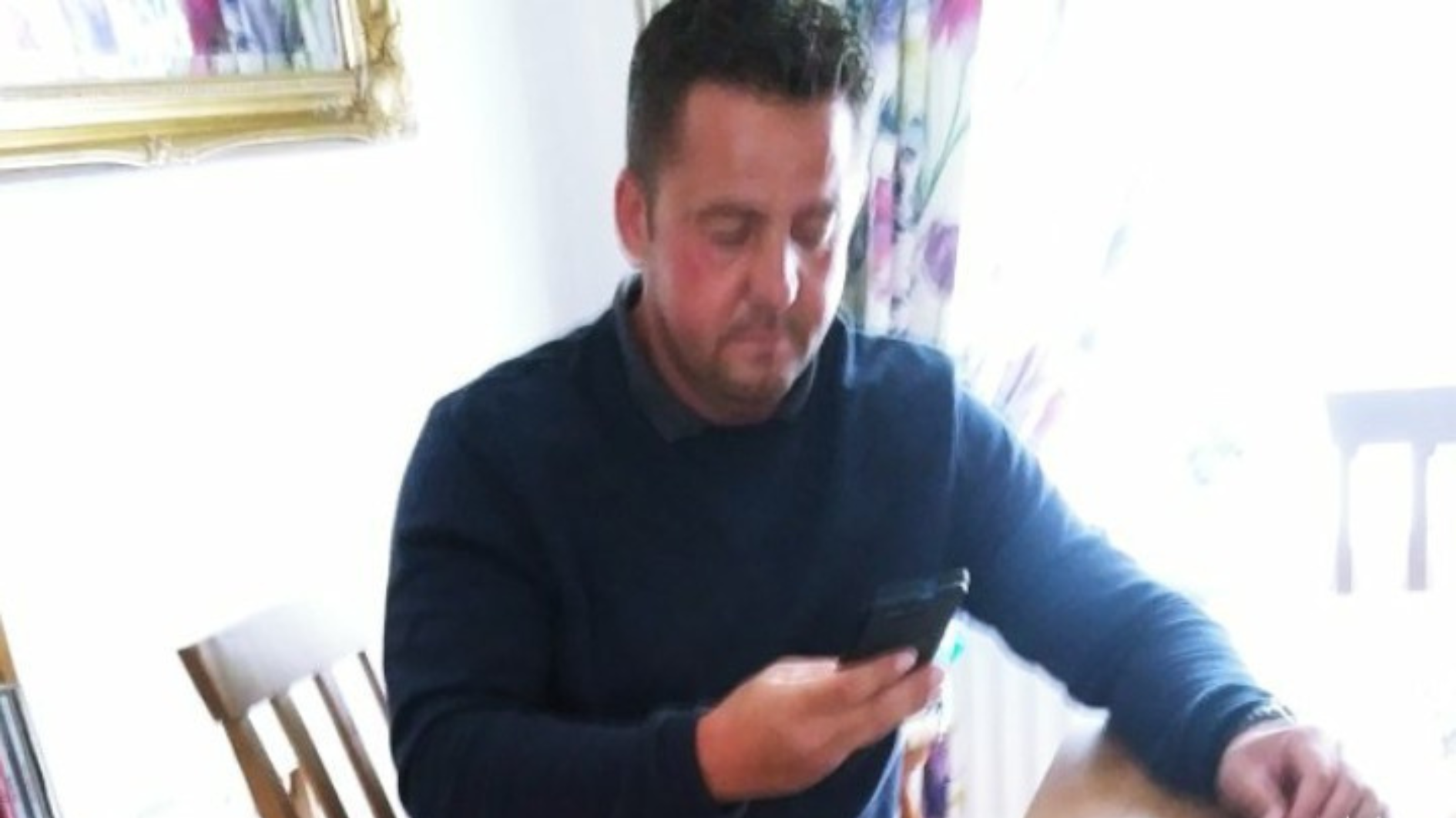 A man sat in a dining room on a chair with arms on the table looking at his phone