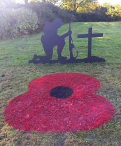 A painted red and black poppy on grass, with a cross and Tommy soldier silhouette frame in black, placed on the grass in a field